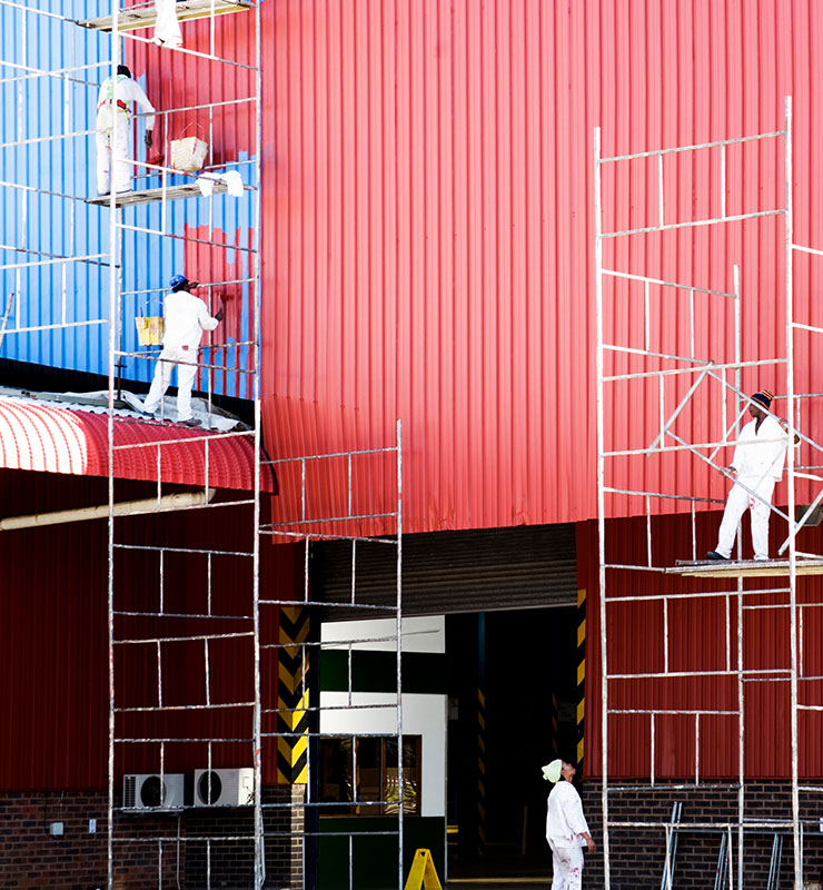 A paint crew stands on a ground scaffold and applies red paint to a metal building exterior